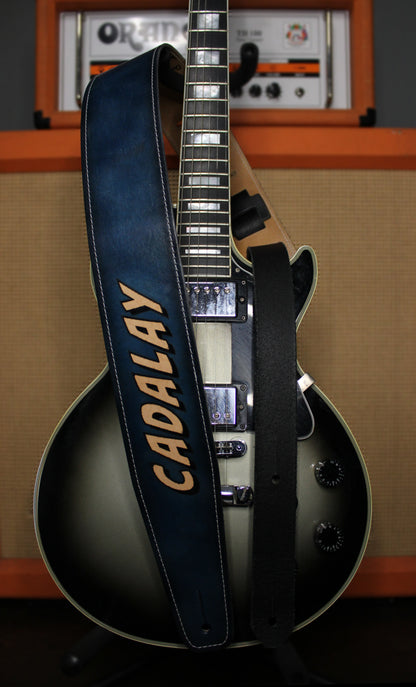 A black and gray sunburst electric guitar with a dark blue leather strap featuring the name 'CADALAY' in beige lettering, set against a warm orange and beige amplifier background.