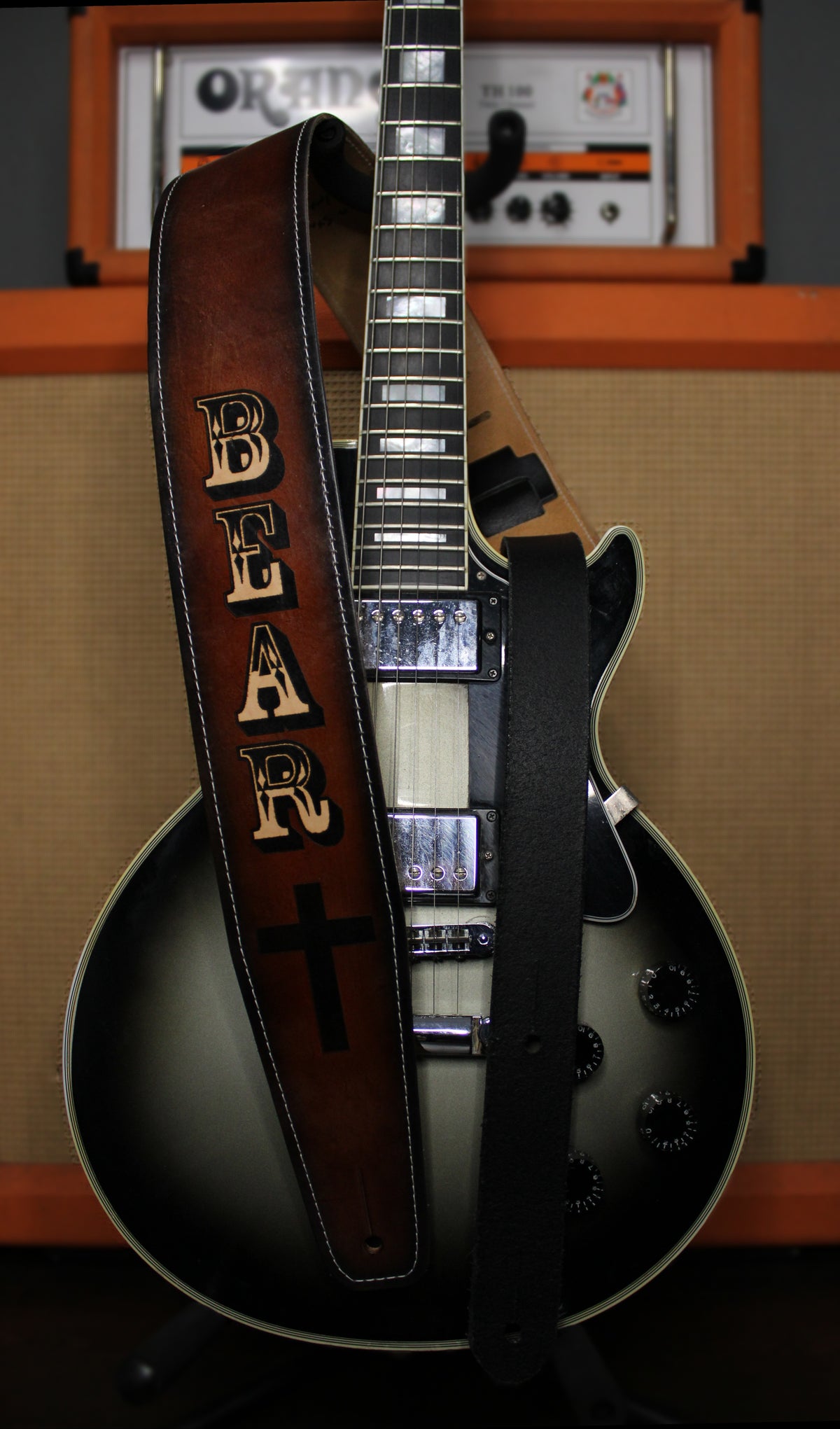A close-up of an electric guitar with a brown leather strap featuring the word 'BEAR' in large black letters and a black cross, set against an orange amplifier.