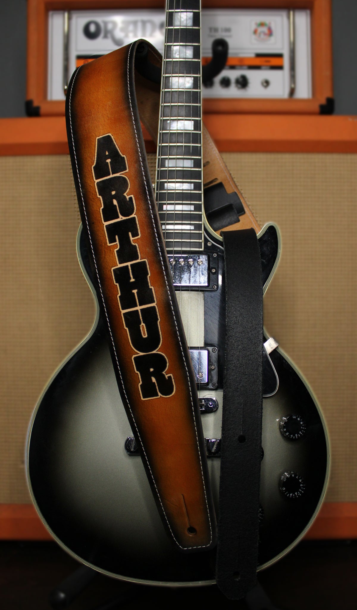 A custom electric guitar strap with the name 'ARTHUR' in black block letters on a brown sunburst leather strap, draped over a black and silver electric guitar in front of an orange amplifier.