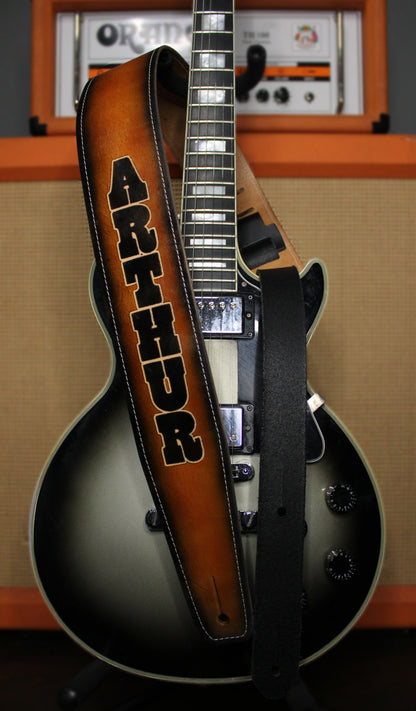 A custom electric guitar strap with the name 'ARTHUR' in black block letters on a brown sunburst leather strap, draped over a black and silver electric guitar in front of an orange amplifier.